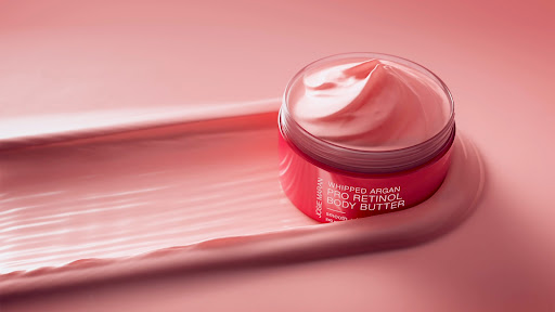 Product shot of Josie Maran Whipped Argan Pro Retinol Body Butter, featuring pink and white swirl of cream on pink background.