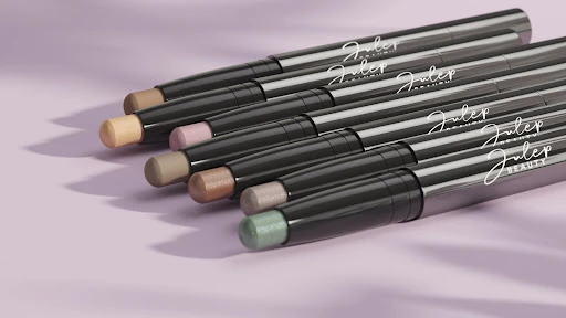 Group of Julep brand eyeshadow sticks in various colors with black packaging, arranged on a lilac surface under soft lighting.