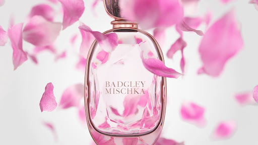 Elegant perfume bottle by Badgley Mischka surrounded by delicate pink rose petals on a soft gradient background in a beauty shot.
