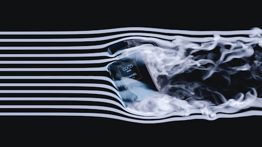 A black Chanel perfume bottle sits in a studio with white smoke, illuminated by studio lighting on a black background.