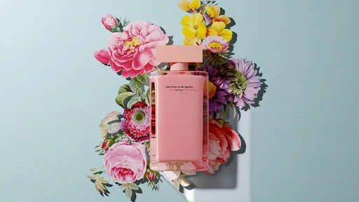 Still life photo of a light pink Narciso Rodriguez perfume bottle, surrounded by an arrangement of colorful flowers on a light blue background.