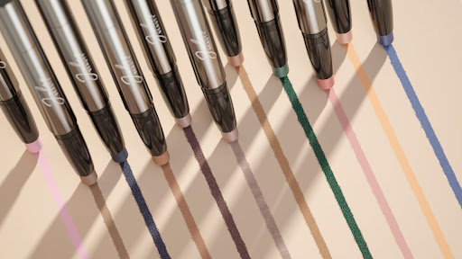 Several silver eyeliner pencils are displayed with the tips marking colourful stripes on a beige background in a studio setting.