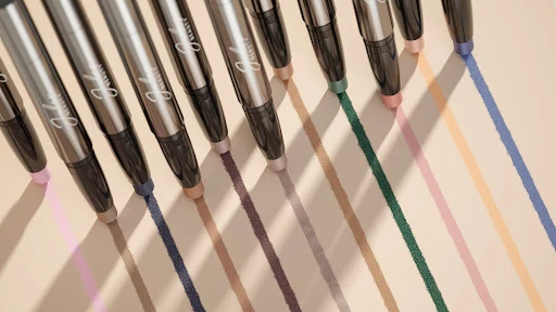 Several silver eyeliner pencils are displayed with the tips marking colourful stripes on a beige background in a studio setting.