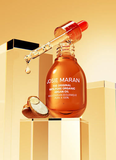 Josie Maran Argan Oil bottle with dropper on clear blocks, featuring oil droplet, product label, and halved argan nut in studio lighting.