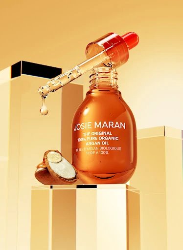 Josie Maran Argan Oil bottle with dropper on clear blocks, featuring oil droplet, product label, and halved argan nut in studio lighting.