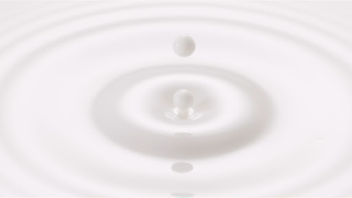 A milk drop falls into a pool of milk, creating circular ripples on the surface of the white liquid in a minimalist food photo.
