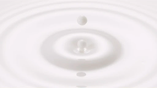 A milk drop falls into a pool of milk, creating circular ripples on the surface of the white liquid in a minimalist food photo.
