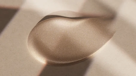 Close-up of a shimmery champagne gold cosmetic cream swatch on a neutral matte surface under soft light.