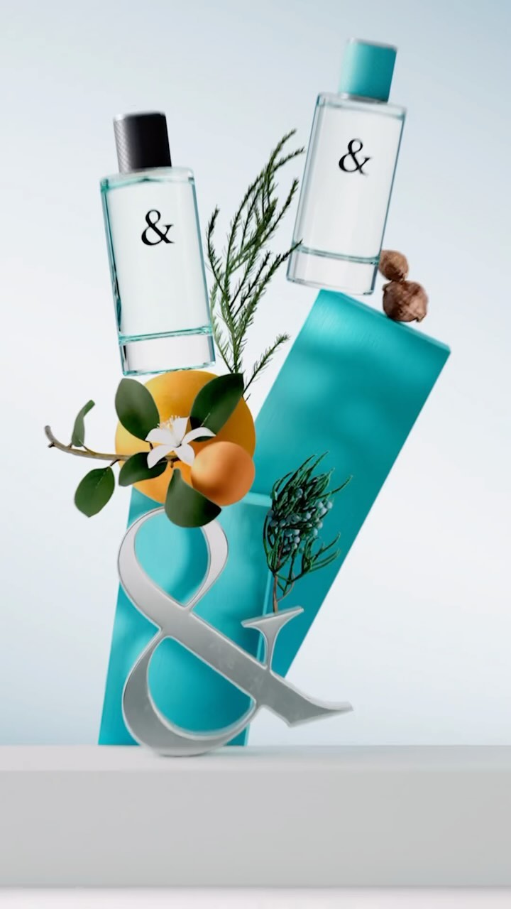 Still life of two perfume bottles featuring a silver '&' symbol, green foliage, and citrus fruit against a light blue background.
