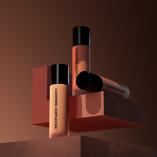 Product shot of three tan-colored foundation bottles with black caps, arranged on geometric blocks against a brown background.