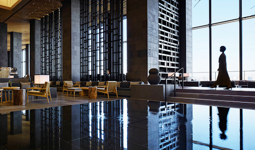 Spacious hotel lobby with high ceilings, lounge areas, a reflecting pool, and a woman walking near large windows.