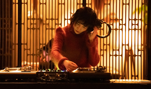 Woman wearing headphones mixing music at a DJ console with turntables in a warm-toned interior with a patterned screen.