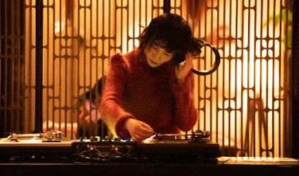 Woman in red turtleneck sweater adjusting DJ equipment with headphones in front of a patterned screen
