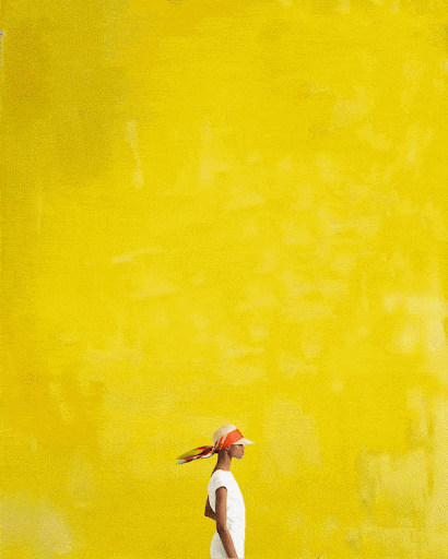 Woman in white dress and headscarf walking against bright yellow painted wall. Side view, full shot, vibrant background.