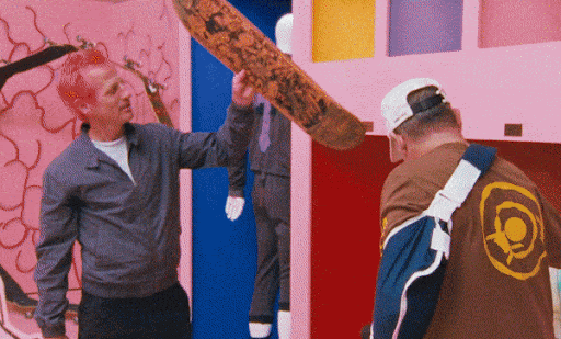 Two men in an art gallery look at a skateboard with pink walls, a mannequin, and colorful geometric wall art in the background.