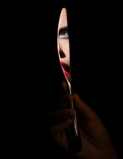 Cropped artistic close-up of a woman's face reflected in a shiny knife blade held by a hand against a stark black background.