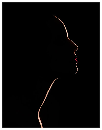 Striking close-up of a backlit woman's silhouette in profile, showcasing her face and neck against a stark black backdrop with red lipstick.