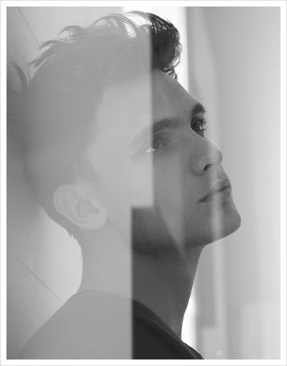 Monochrome portrait of a dark-haired man, gazing upward, with a partially obscured face and a soft, diffused background light.