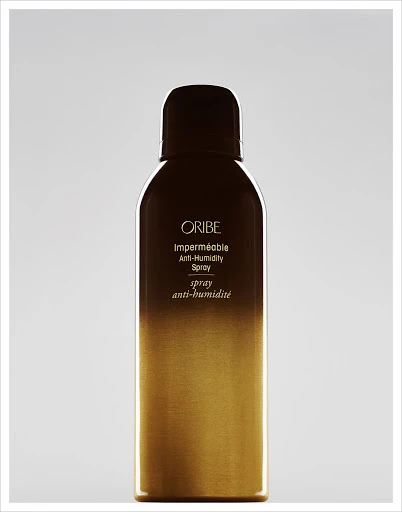 Oribe Impermeable Anti-Humidity Spray bottle with a brown top fading into a gold bottom on a neutral grey background.