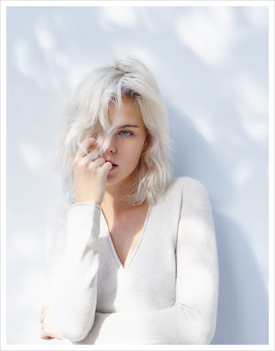 Portrait of a young adult female with platinum blonde hair, wearing a white long-sleeve shirt, against a light blue and white backdrop.