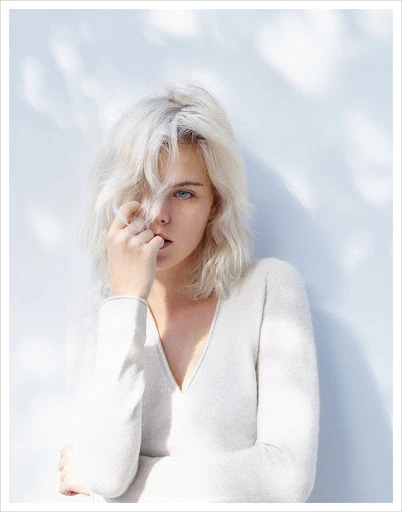 Portrait of a young adult female with platinum blonde hair, wearing a white long-sleeve shirt, against a light blue and white backdrop.