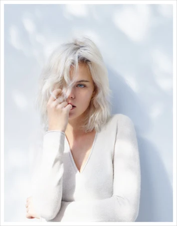 Portrait of a young adult female with platinum blonde hair, wearing a white long-sleeve shirt, against a light blue and white backdrop.