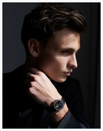 Portrait of an adult male wearing a black suit and a wristwatch looking off into the distance in a dimly lit indoor setting.