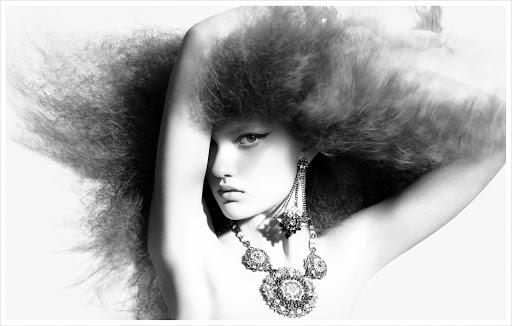 Black and white portrait of a woman with voluminous textured hair, winged eyeliner, and an ornate statement necklace.