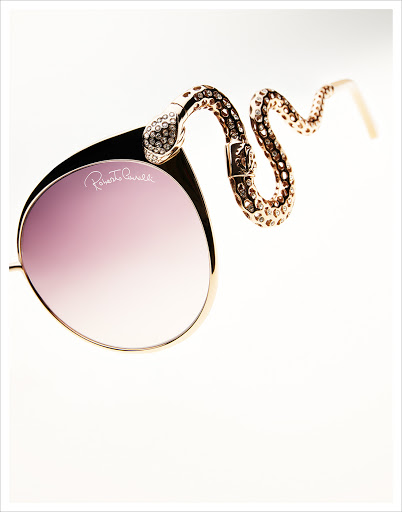 Elegant gold-framed sunglasses with purple lenses feature a bejeweled, snake-shaped arm, presented on a bright white background in a studio shot.