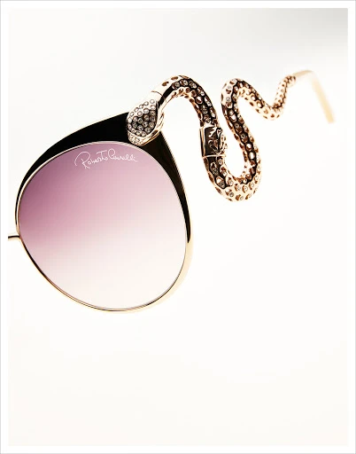 Elegant gold-framed sunglasses with purple lenses feature a bejeweled, snake-shaped arm, presented on a bright white background in a studio shot.