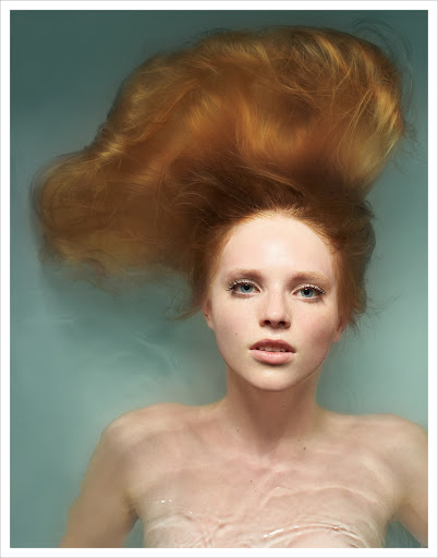 Portrait of a young red-haired woman immersed in water, captured in a close-up studio shot with soft, diffused lighting.