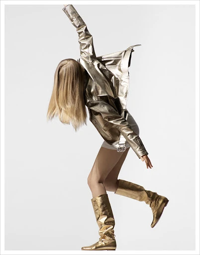 Fashionable woman strikes a dynamic pose in gold metallic jacket, shorts, and high boots against a clean white studio backdrop.