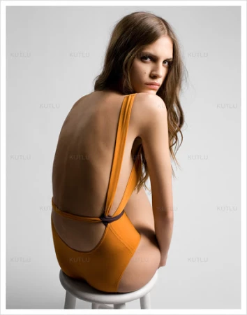 Model wearing a mustard yellow swimsuit sitting on a white stool against a light background, photographed by Kutlu.