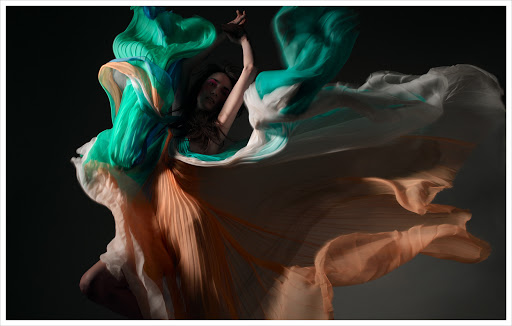 Artistic portrait of a person draped in flowing teal, white, and peach fabric against a dark background in an expressive pose.