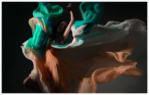 Artistic portrait of a person draped in flowing teal, white, and peach fabric against a dark background in an expressive pose.