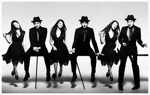 Black-and-white studio shot features six adults (four women, two men) sitting and standing in formal wear—hats, suits, and dresses.