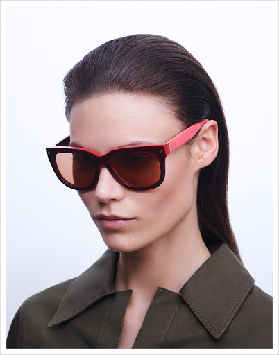 Fashionable woman with dark hair and olive green shirt wearing dark sunglasses with coral accents against a white background.