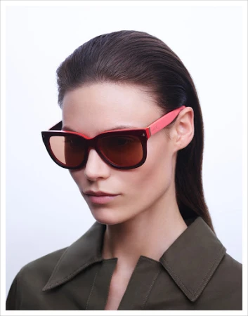 Model wearing dark tinted sunglasses with pink temples and an olive green collared shirt against a white backdrop.