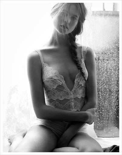 Monochrome portrait of a young adult woman in a lace bodysuit, with a braided hairstyle, posing in an indoor setting with soft lighting.