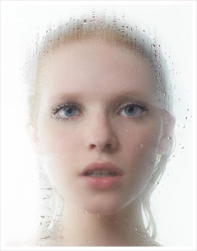 Close-up portrait of a fair-skinned woman with blue eyes looking through a wet glass surface, soft lighting, white background.
