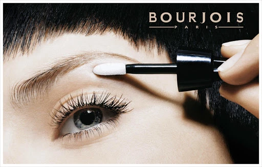 Close-up of a woman's face with makeup application, featuring an eye with long lashes and a cosmetic tool applying shadow to the eyebrow.