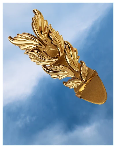 Elevated view of a single golden sandal with ornate wing details against a blue sky with wispy clouds for fashion or product advertising.