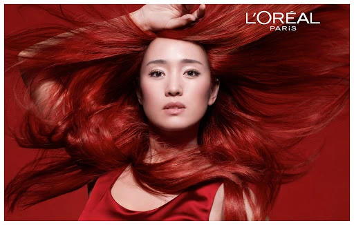 Beauty shot of an Asian woman with voluminous red hair and a red dress posing in front of a solid red background, featuring L'Oreal Paris logo.