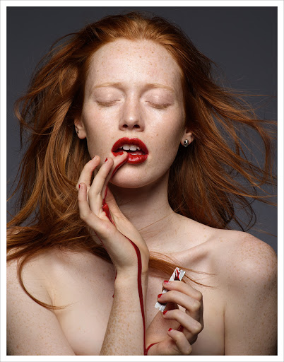 Close-up portrait of a red-haired woman with freckles, red lips, and red pigment dripping down her hand photographed by Kutlu.