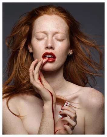 Close-up portrait of a red-haired woman with freckles, red lips, and red pigment dripping down her hand photographed by Kutlu.
