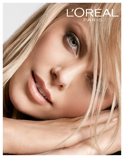 Close-up portrait of a blonde woman with fair skin and green eyes. She is posed with her hands crossed beneath her face. 'L'Oreal Paris' logo visible.