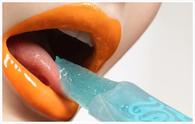 Close-up macro shot of lips painted bright orange with a blue gel popsicle touching the tongue against a white background
