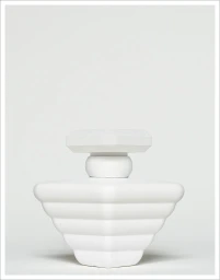 A white sculptural perfume bottle with geometric tiered ridges set against a plain light grey backdrop.