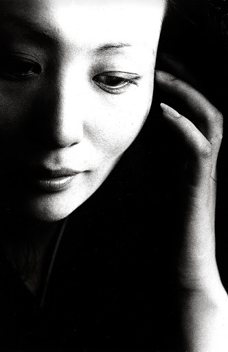 Monochrome portrait of an Asian woman with her hand near her face, set against a dark background in soft, diffused lighting.