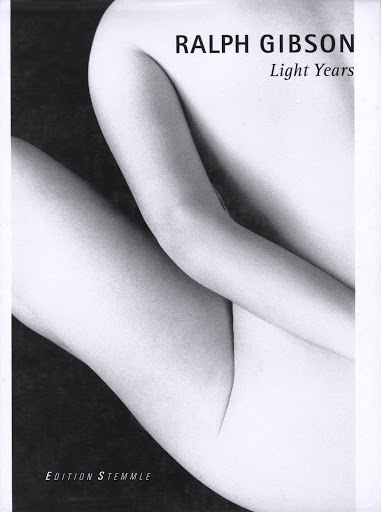 Monochrome photography featuring a close-up of a partially visible nude adult female figure, in a seated pose with black background and text overlay.